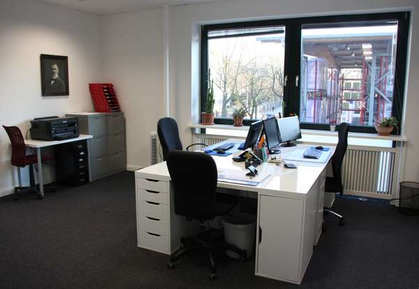 Photo of an office room with 2 windows, in front of it a large desk with 3 workstations, on the left filing cabinets and a printer.